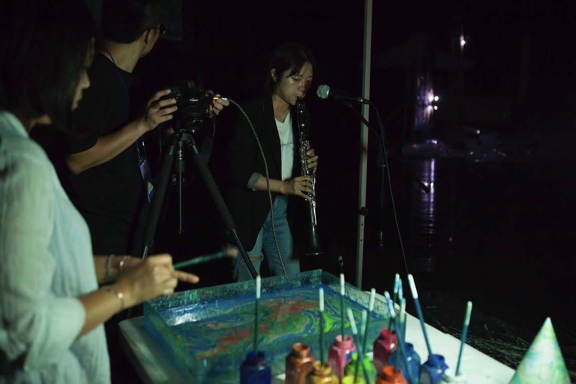 At night by the water, Lee plays clarinet into a microphone while a marbling artist works pigment across a tray and a camera films it.