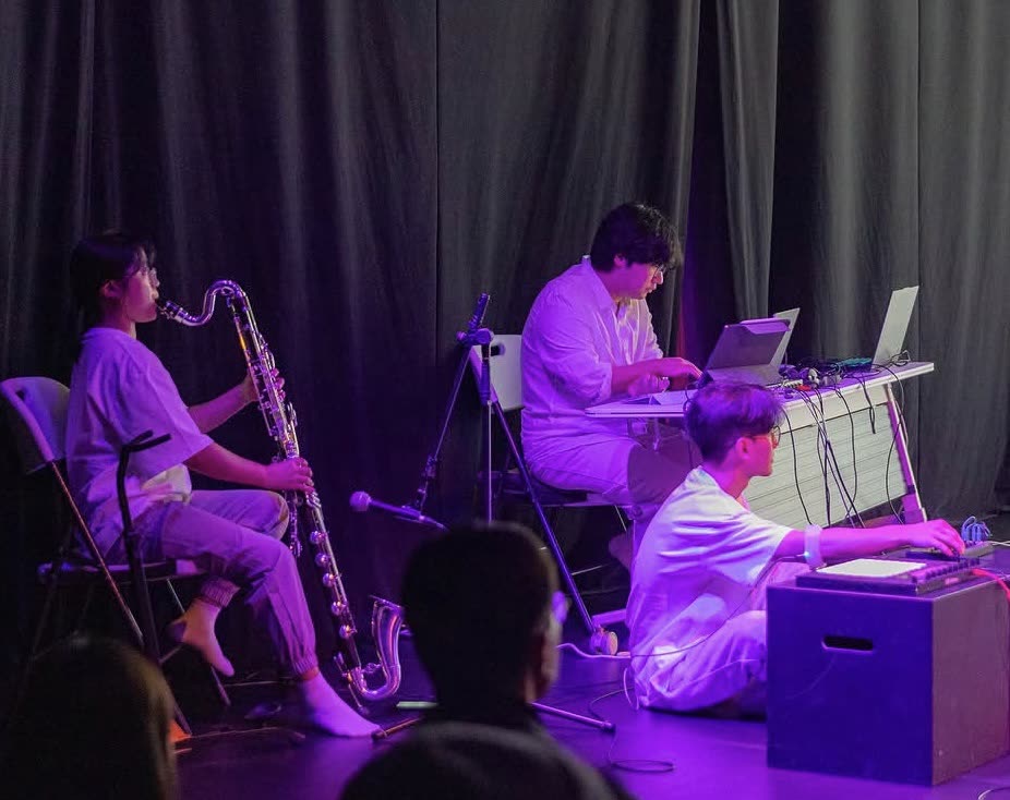 Lee plays bass clarinet under violet stage light while two electronic musicians work at laptops and a controller.